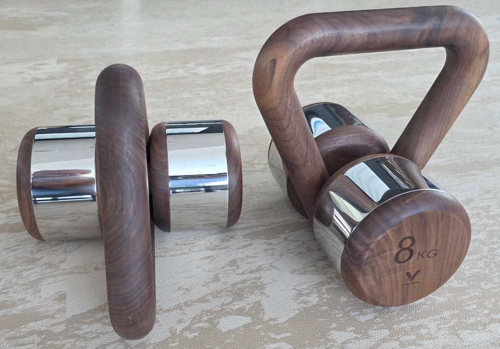 Two wooden dumbbells with polished chrome ends and a wooden kettlebell handle, displayed on a light beige textured surface.