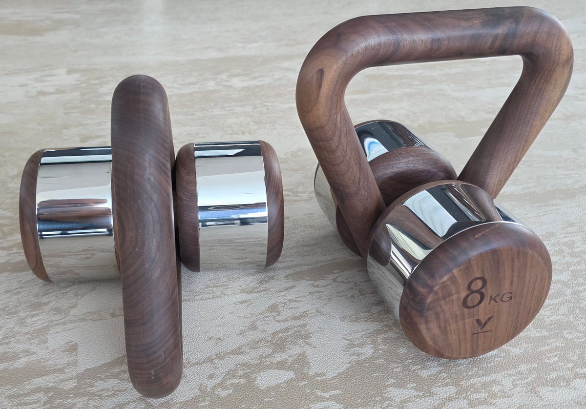 Two wooden dumbbells with polished chrome ends and a wooden kettlebell handle, displayed on a light beige textured surface.
