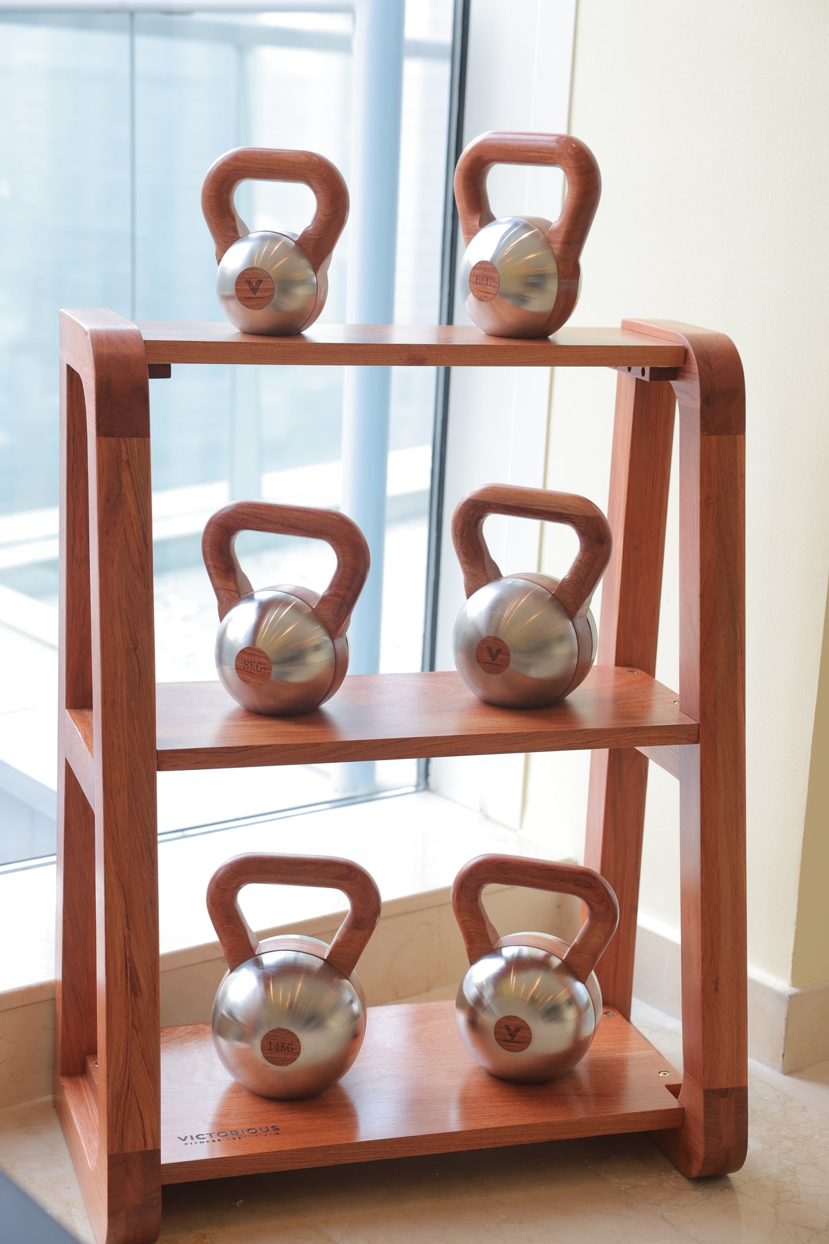 A three-tier wooden kettlebell rack holding six stainless steel kettlebells with brown handles, positioned indoors near a window.